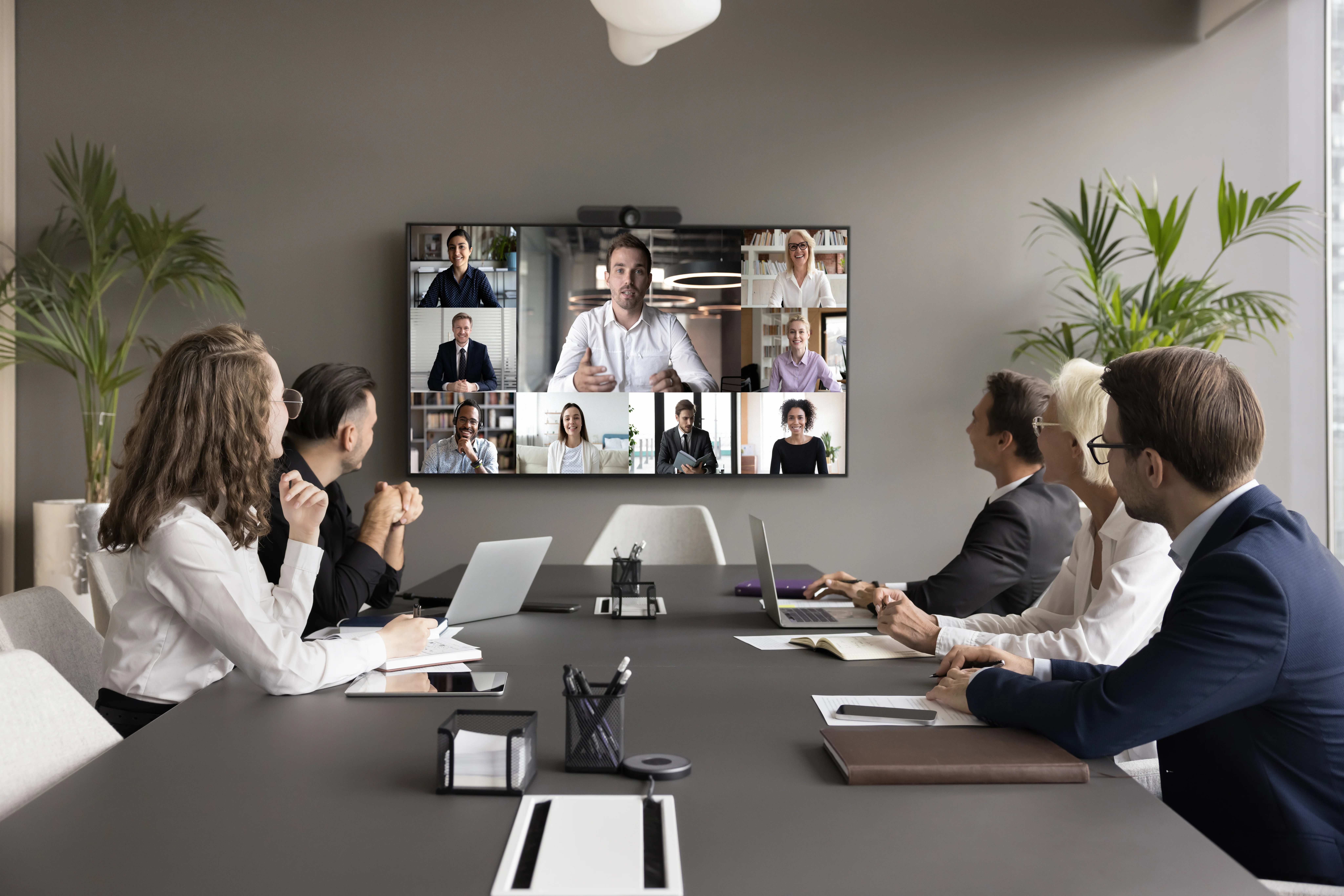 a work team gathers in a conference room to hold a virtual meeting; remote team members are displayed on the large screen at the front of the room.